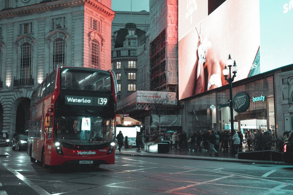 A busy urban street scene in Waterloo with a prominent red double-decker bus displaying route number 139 and destination WaterLoo moving through the foreground. Behind the bus, there are several pedestrians walking along the pavement, some gathered near an entrance to a commercial building with large glass windows and signage including a 'Boots' beauty store. To the right, a classic street lamp with multiple lanterns is visible, partially obscured by a large digital billboard that shows abstract marketing imagery with human legs and a hand. The surrounding architecture includes historic and modern buildings, with the historic building on the left featuring arched windows and decorative stonework, while the modern structures on the right incorporate glass facades and scaffolding. The scene is illuminated by ambient street lighting and the glow from the digital billboard, reflecting an active city environment that contextualizes private transport and pedestrian activity, relevant to discussions of alternative waste removal methods and urban rubbish management by companies like Waste Collection Waterloo.