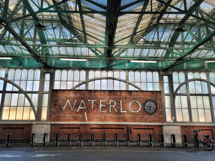 Interior view of a transportation hub with a large brick wall prominently displaying the word 'WATERLOO' in metal letters, accompanied by a circular emblem. The structure features extensive iron framework supporting a glass and metal roof, allowing natural light to filter in. The brick wall with the signage is positioned above a paved area that includes bike racks and bicycles on the right side, suggesting a commuter or public transport environment. The scene is clean and well-maintained, with the industrial design elements and lighting highlighting the architectural details. The setting reflects an urban location that may also serve as a site for managing or handling waste and rubbish collection, hinting at potential alternatives to local authority disposal methods associated with private waste management services like Waste Collection Waterloo, which specializes in rubbish removal solutions.