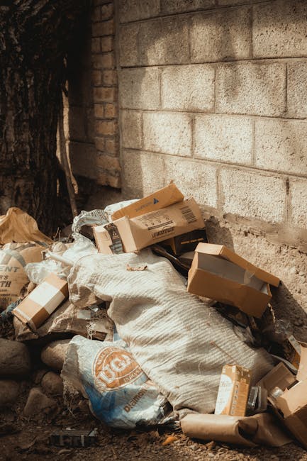 A pile of domestic waste including flattened cardboard boxes, paper bags, and plastic packaging rests against a brick wall and concrete block wall, with some items partially covered by a textured fabric sack. The waste material is situated outdoors on a mixed surface of dirt and small stones, with a large tree trunk visible to the left. The scene appears to depict unmanaged rubbish that could be the subject of private rubbish removal or alternative waste handling services like those offered by Waste Collection Waterloo. Natural daylight illuminates the area, casting minimal shadows. The collection of cardboard and general waste shows typical materials that may be collected for disposal or recycling through independent rubbish clearance services.