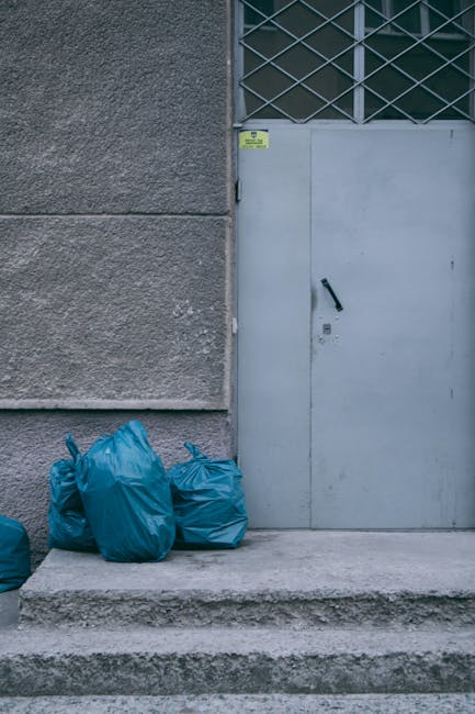 The image shows four blue rubbish bags filled with waste, placed on a concrete step outside a building. The bags are made of lightweight plastic with visible creases and tied openings. They are positioned near a grey metal door with a black handle, set into a white frame, adjacent to a section of rough, textured grey concrete wall. Above the door, there is a rectangular metal security grille with a crisscross pattern, covering a window. The environment appears to be an urban or commercial setting, with the door and wall suggesting an exterior of a warehouse, shop, or industrial space. The lighting is natural, indicating daytime, with no additional equipment or objects present. This scene may reflect a situation of private waste disposal or on-site rubbish collection, which Waste Collection Waterloo could potentially assist with, aligning with alternative waste management options outside standard local authority kerbside collections.