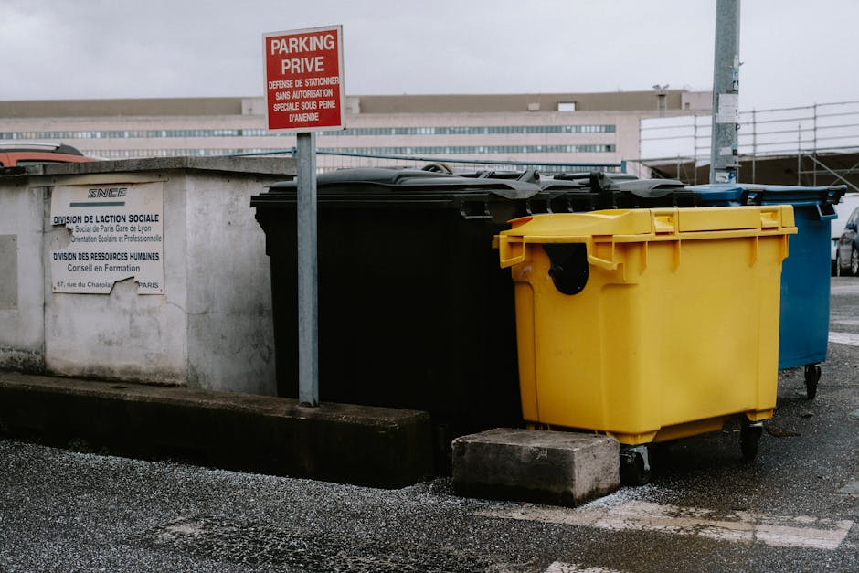 In a public parking area, three large wheeled rubbish bins are positioned next to a concrete wall. The first bin, yellow in color, has a smooth plastic surface with a black lid and is situated closest to the camera on the right. Behind it, a black bin with a slightly textured surface and a matching black lid is aligned, while a blue bin with a similar finish is partially visible further back. The bins are placed on an asphalt surface, slightly elevated on small concrete blocks to facilitate waste collection. A metal pole with a red and white sign indicating 'Parking Privé' is mounted nearby, and a white sign with French text is affixed to the concrete wall behind the bins. The surrounding environment appears to be an urban parking lot, with modern buildings visible in the background under overcast lighting. The scene reflects a typical setup for waste disposal in a commercial or multi-use development, with Waste Collection Waterloo providing a clear example of private waste management practices outside formal municipal collection routes.