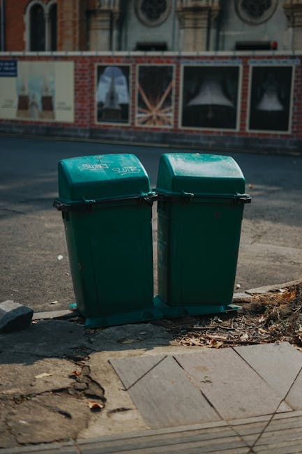 In a public parking area, three large wheeled rubbish bins are positioned next to a concrete wall. The first bin, yellow in color, has a smooth plastic surface with a black lid and is situated closest to the camera on the right. Behind it, a black bin with a slightly textured surface and a matching black lid is aligned, while a blue bin with a similar finish is partially visible further back. The bins are placed on an asphalt surface, slightly elevated on small concrete blocks to facilitate waste collection. A metal pole with a red and white sign indicating 'Parking Privé' is mounted nearby, and a white sign with French text is affixed to the concrete wall behind the bins. The surrounding environment appears to be an urban parking lot, with modern buildings visible in the background under overcast lighting. The scene reflects a typical setup for waste disposal in a commercial or multi-use development, with Waste Collection Waterloo providing a clear example of private waste management practices outside formal municipal collection routes.