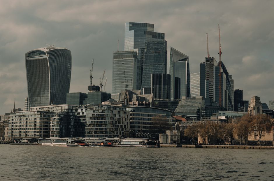 A panoramic view of a modern city skyline featuring several tall glass-clad skyscrapers with reflective surfaces, including a distinctive curved tower on the left and multiple high-rise buildings under construction with visible cranes on their rooftops. In the foreground, low-rise residential and commercial buildings with balconies and flat roofs are situated along the waterfront, with some boats docked or sailing nearby on a calm river. The sky above is overcast with grey clouds, creating a muted lighting across the scene. This urban environment, characteristic of central London's financial district, showcases a dense concentration of contemporary architecture that might be targeted for private waste collection and on-site clearance services by companies such as Waste Collection Waterloo, emphasizing the importance of alternative rubbish removal solutions for large-scale city developments.