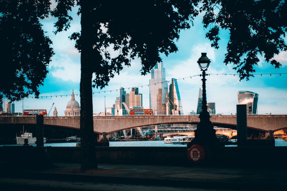 A scenic view of the London skyline seen through a park with two large trees in the foreground, their dark silhouettes framing the scene. A vintage-style street lamp is positioned centrally, with a string of decorative lights stretching across the image above the walkway. The skyline features prominent modern skyscrapers, including the Shard and the Walkie Talkie building, along with historic structures like St. Paul's Cathedral with its large dome, all set against a partly cloudy sky with patches of blue. Below the buildings, a river with several boats and vessels is visible, with a bridge crossing over it. The scene captures an urban landscape that could be relevant to private waste collection or on-site clearance activities, as part of the varied methods of waste handling in the city, with the surroundings suggesting a public outdoor area near the water and city centre. The lighting indicates late afternoon or early evening, casting a natural glow over the scene, contributing to the overall professional and neutral tone suitable for a waste removal service context.